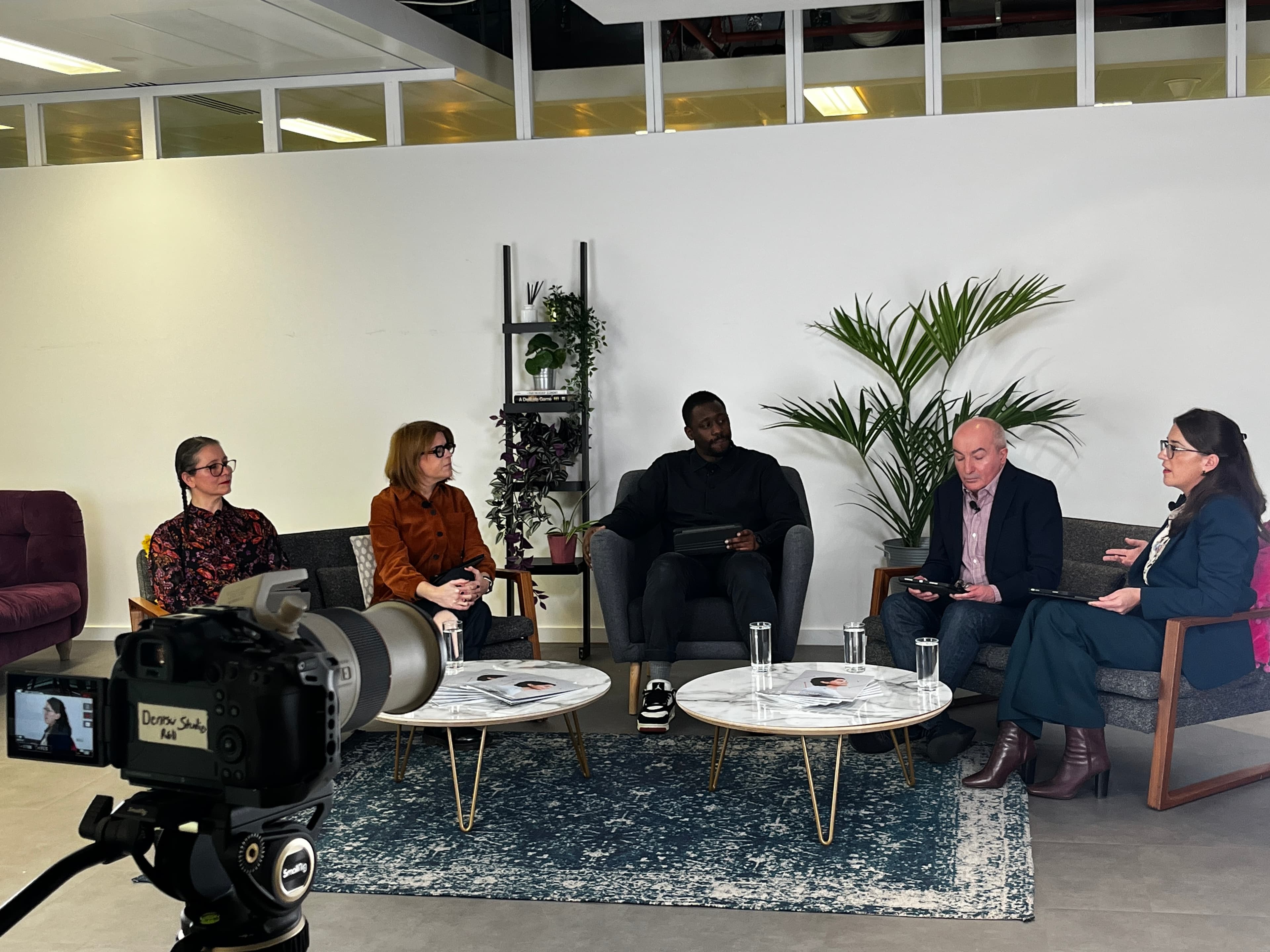 A group of five people standing together indoors, smiling at the camera. Two individuals on the left and one on the right are holding printed copies of a report featuring a person wearing headphones on the cover. They are dressed in smart and semi-formal clothing, standing in a modern studio-style setting with a rug, chairs, and soft lighting in the background.