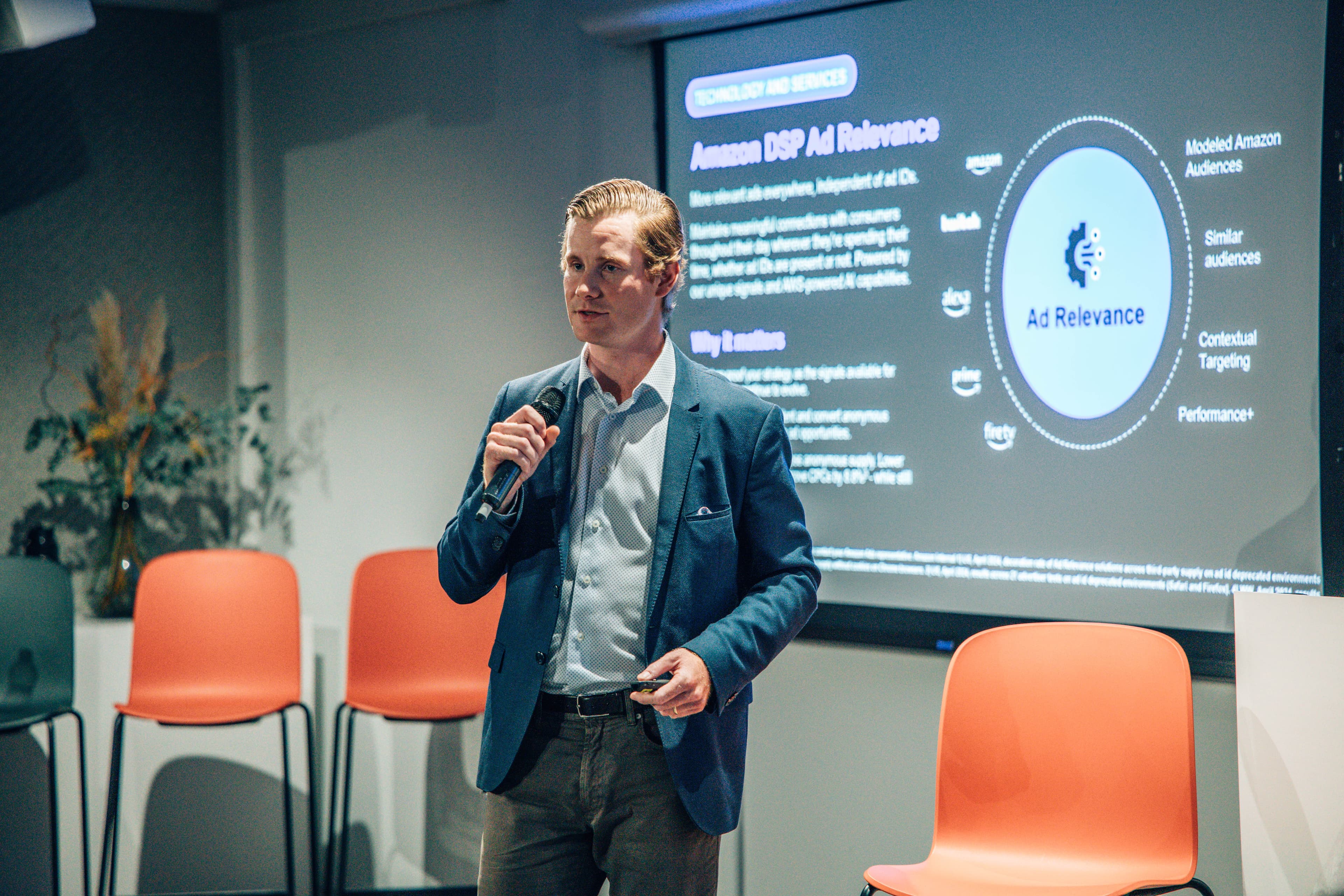 man presenting in front of screen with writing on it and orange chairs in the background