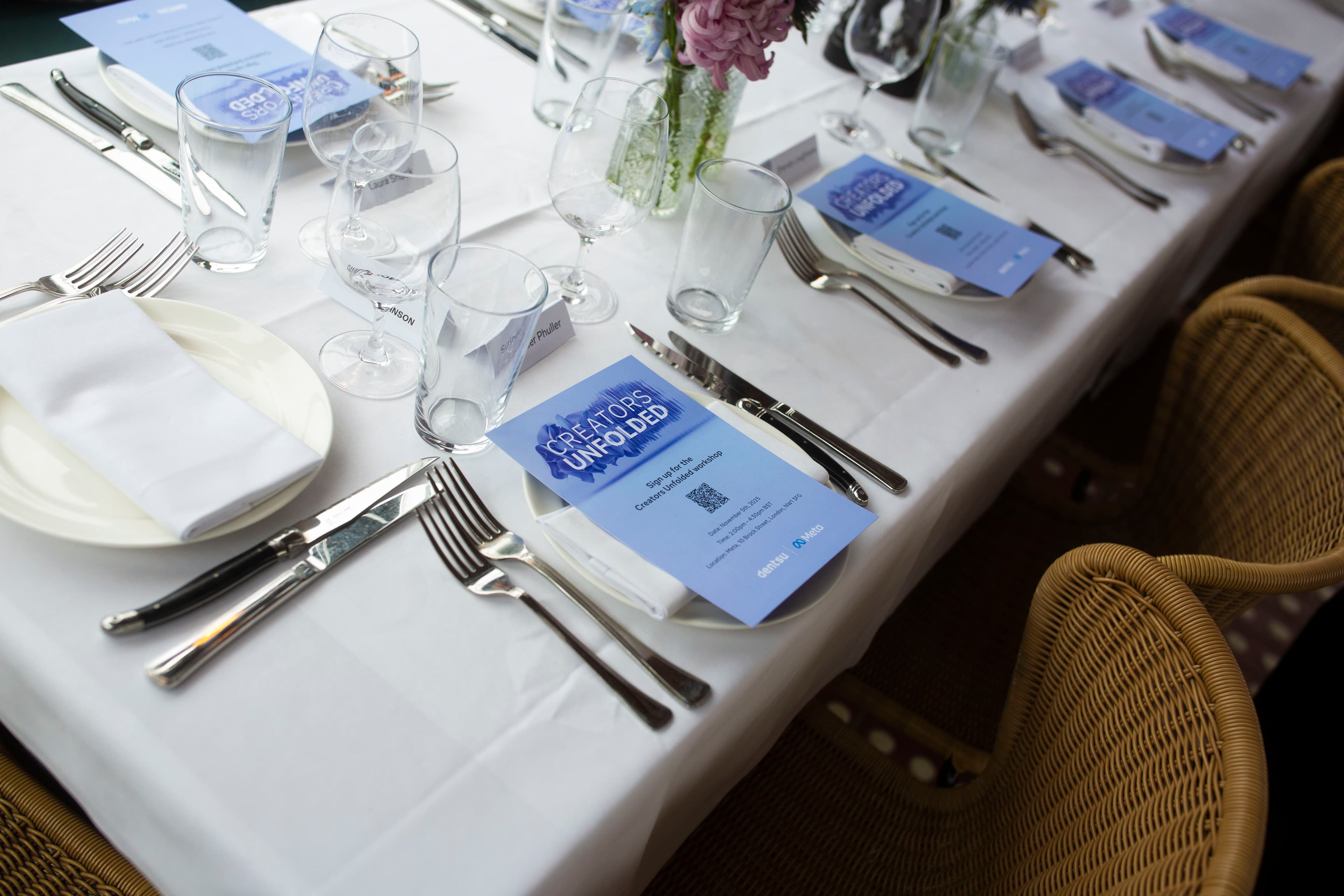 A neatly arranged dining table set for the dentsu x Meta “Creators Unfolded” event, featuring branded menus, glassware, and floral centrepieces prepared for guests